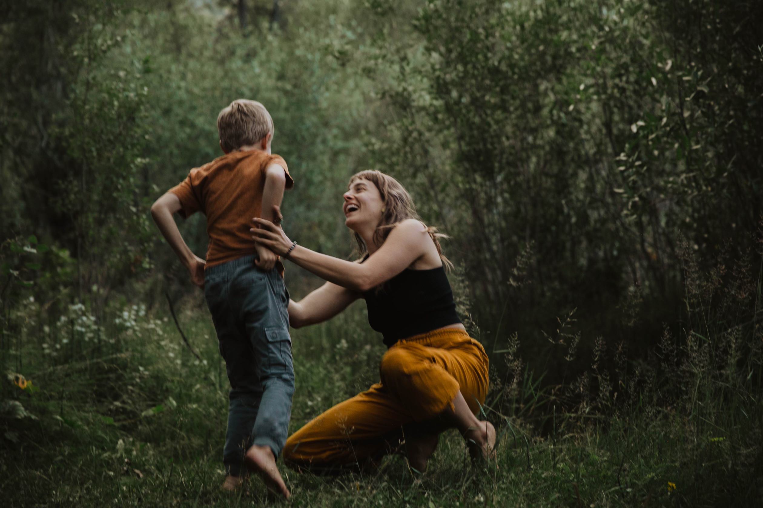 a mother holding her sons hands in the forest. they are smiling and laughing together. 