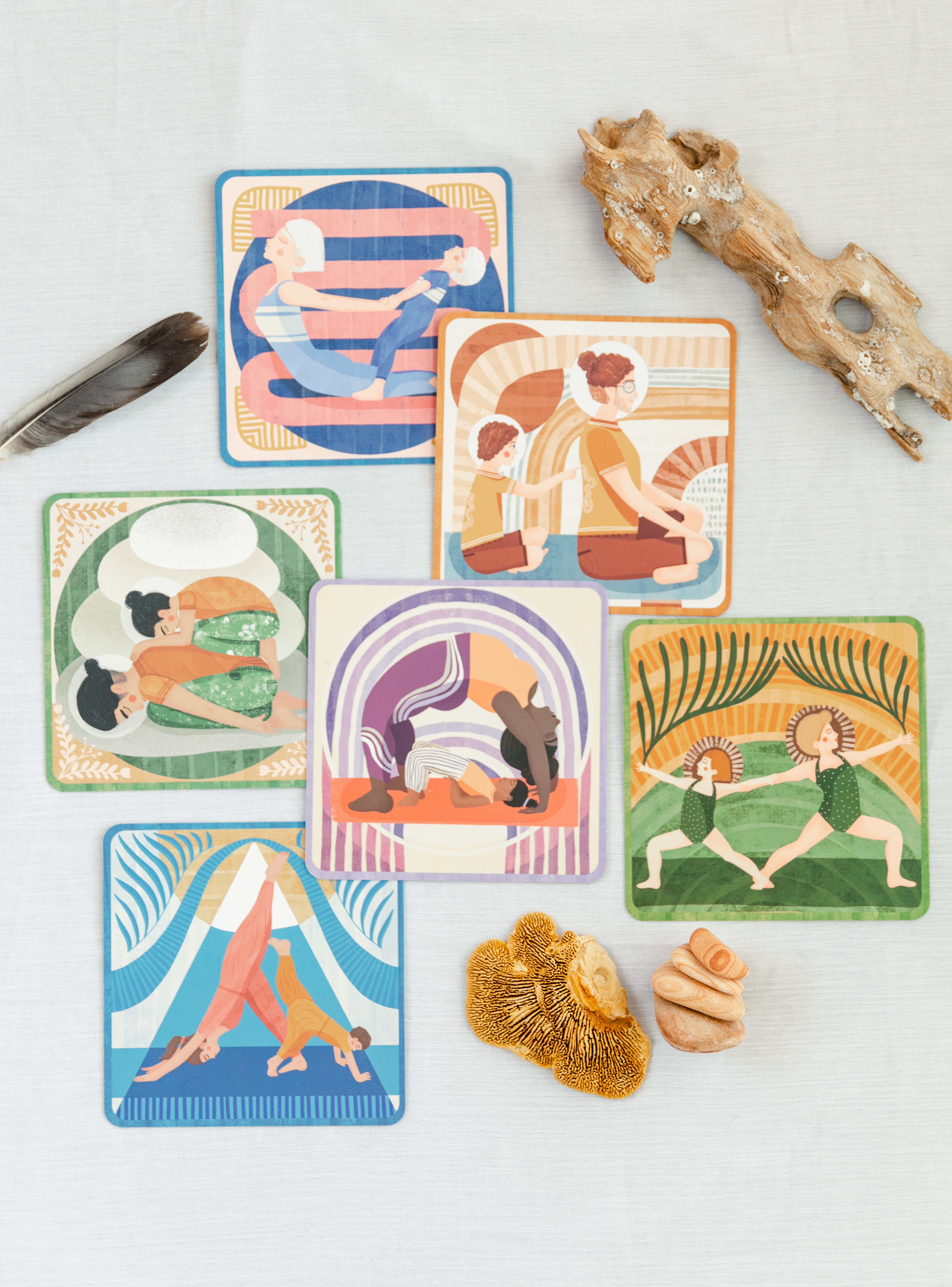 a series of family yoga partner pose cards with diverse families represented. each are a different color relevant to the element they represent. blue is for water, green for earth, orange for fire, and purple for air. The cards are artistically overlaid on top of one another and surrounded by nature items to bring them together. the cards all show a parent and child doing a pose together. each is different and unique. 