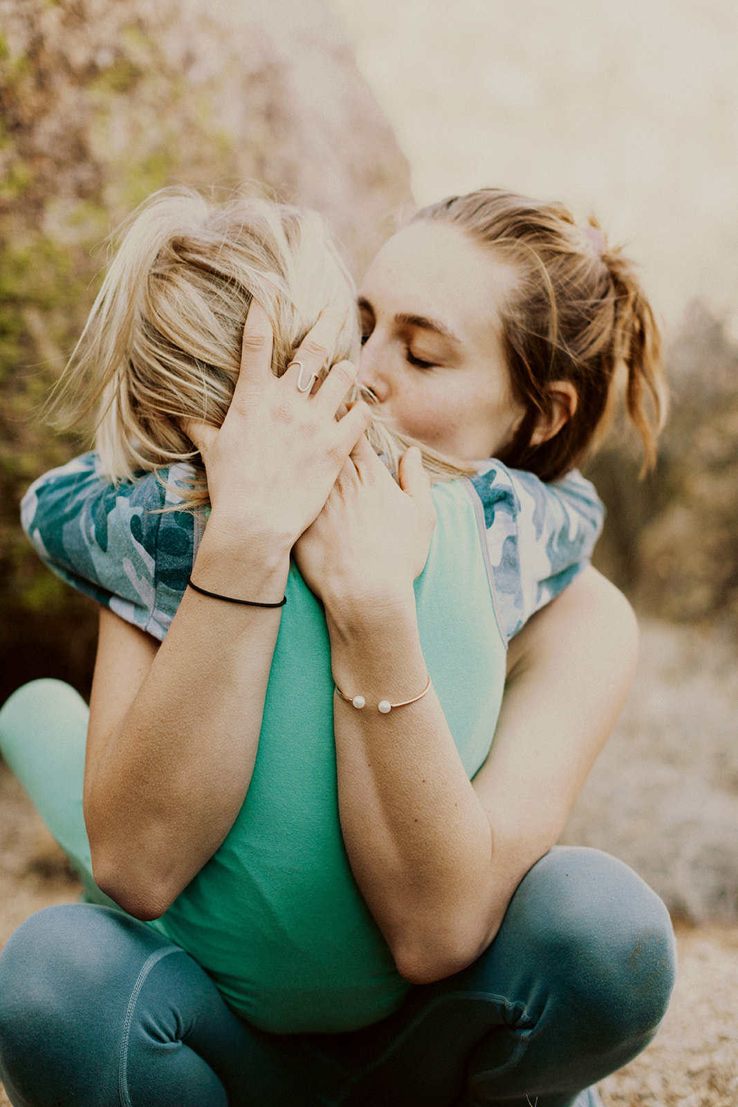 A parent holding her son out in a rocky landscape giving her child a gentle kiss on the cheek while soaking in the love and connection they have for one another. 